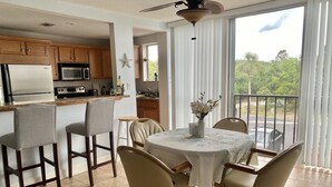 A modern kitchen with wooden cabinets, a refrigerator, microwave, and stove. There is a dining area with a table, chairs, and a vase of flowers. A ceiling fan is visible above the table. - Long term Travelers: spacious 2 BR beach condo (Ormond Beach)