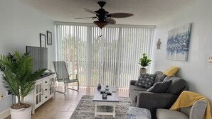 A living room with a sofa, ottoman, coffee table, and a TV on a cabinet. There is a ceiling fan, a window with blinds, and a plant. - Long term Travelers: spacious 2 BR beach condo (Ormond Beach)