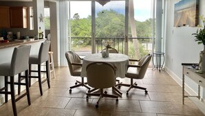 A spacious dining area with a round table and chairs, large windows with a view of greenery, and a ceiling fan. - Long term Travelers: spacious 2 BR beach condo (Ormond Beach)