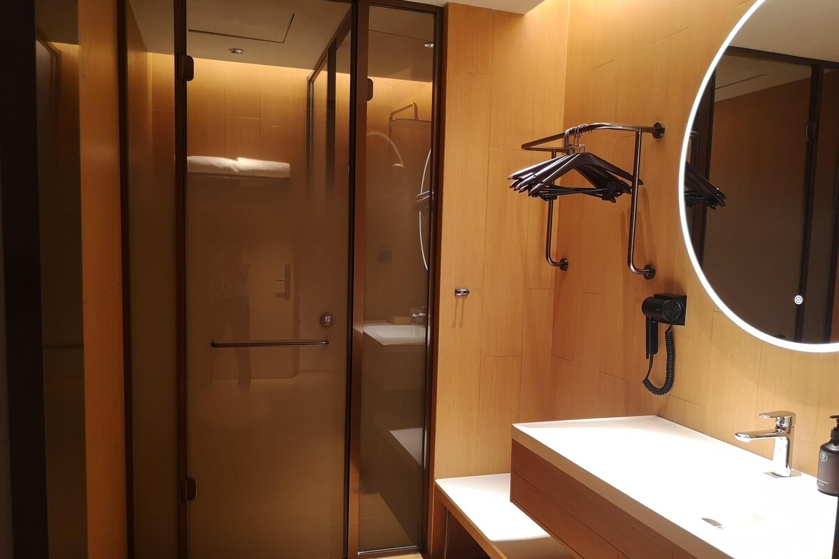 A modern bathroom with a glass shower enclosure, a wooden vanity, a round mirror, and a hairdryer mounted on the wall.