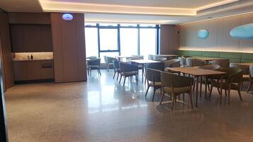 A modern dining area with tables and chairs, a kitchen area with cabinets, and large windows allowing natural light.