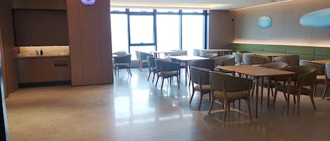 A modern dining area with tables and chairs, a kitchen area with cabinets, and large windows allowing natural light.
