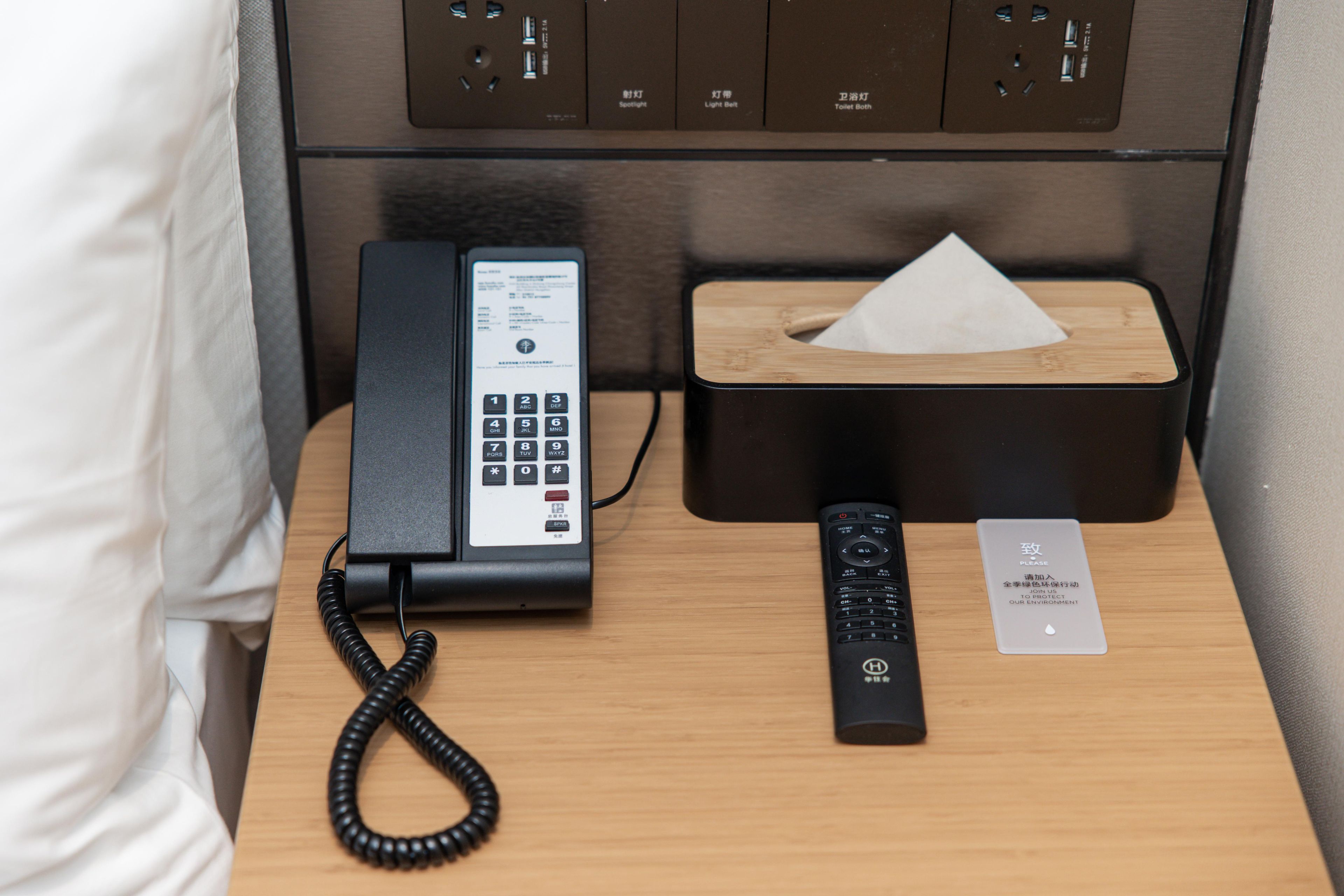 A hotel room with a telephone, a remote control, and a tissue box on a wooden nightstand.