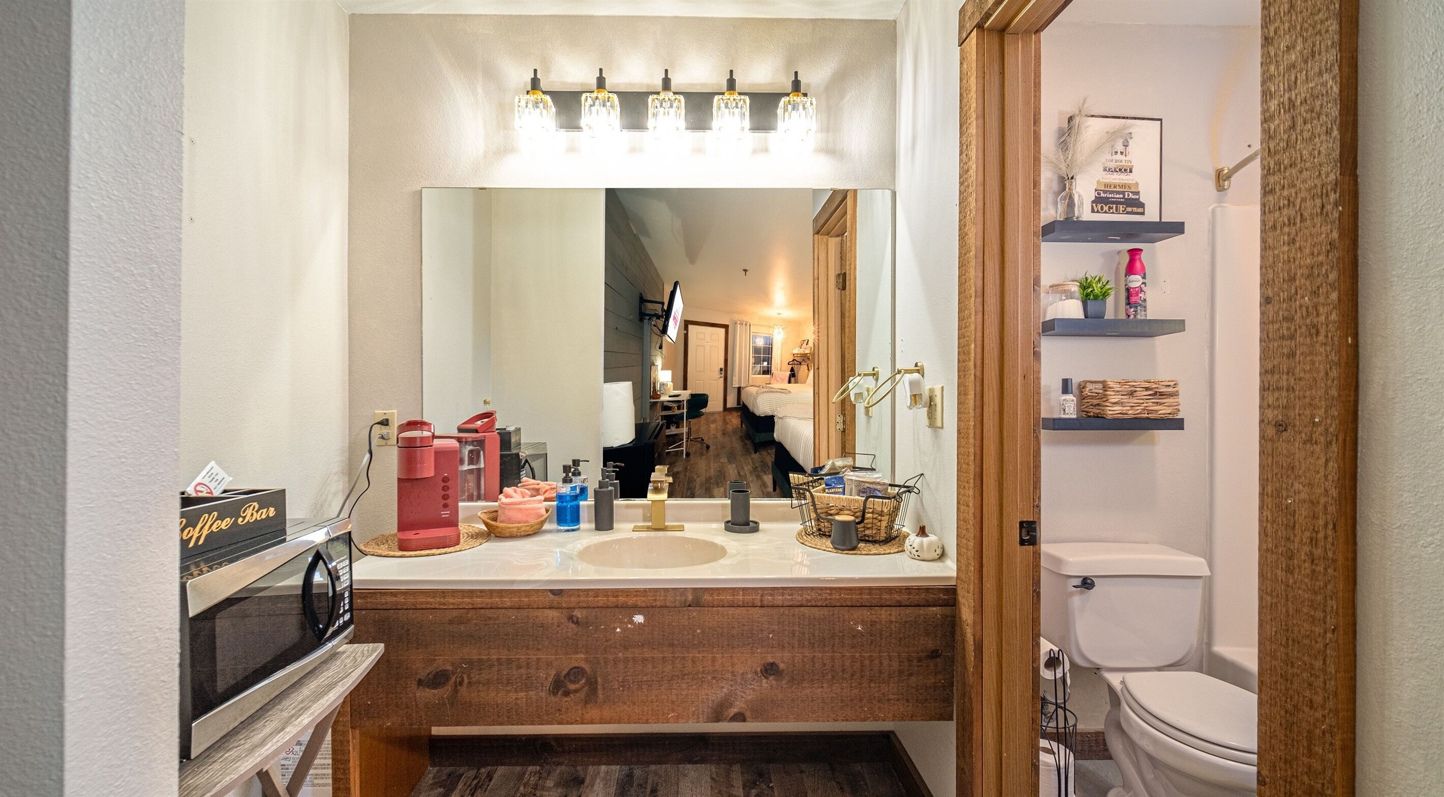 A bathroom with a wooden vanity, a sink, a mirror, and a toilet. There are shelves with toiletries and a coffee machine on the vanity.