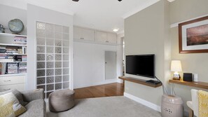 A modern living room with a flat-screen TV, a bookshelf, a round ottoman, and a painting on the wall.