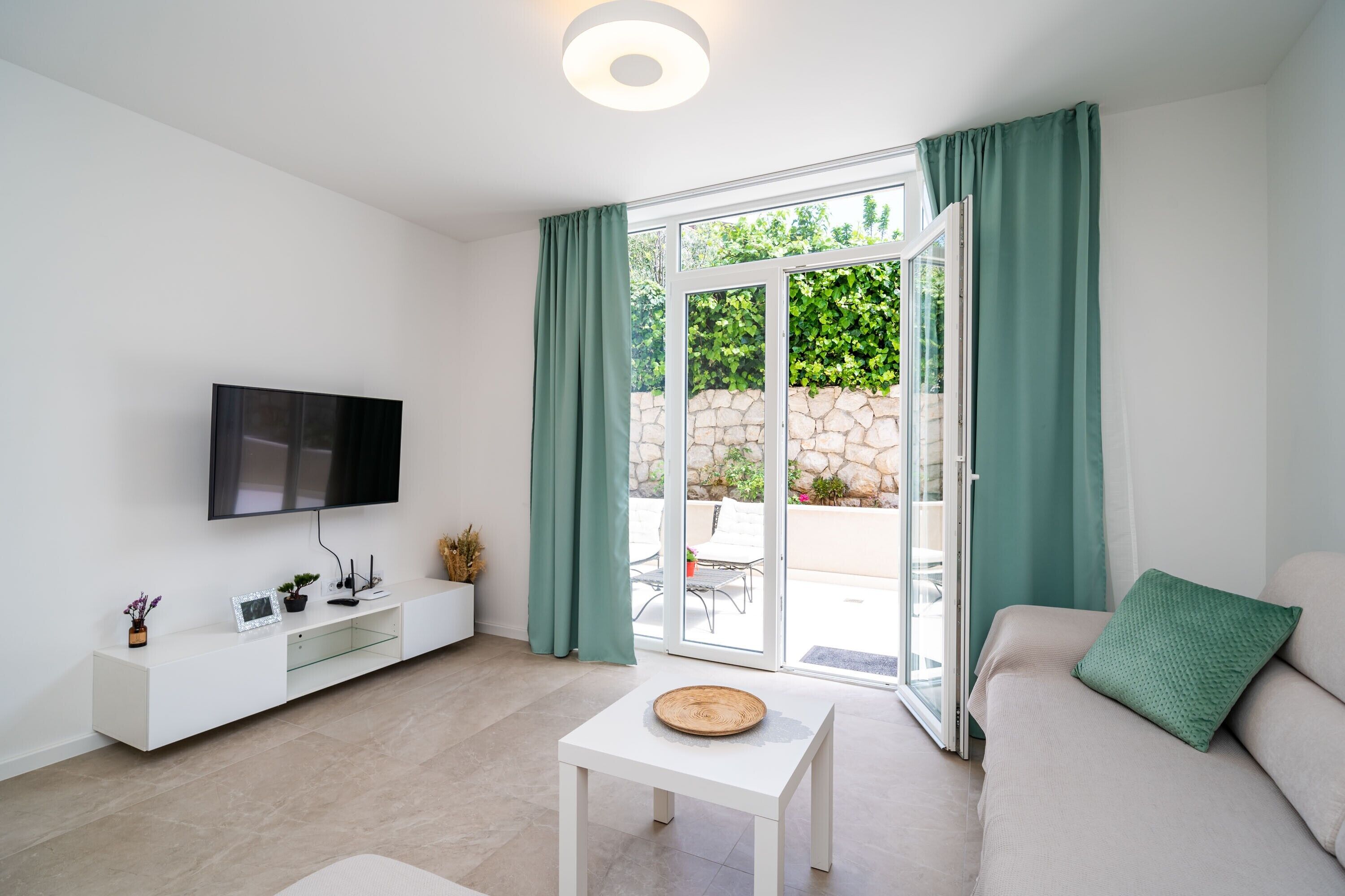 A modern living room with a flat-screen TV, a white coffee table, a sofa with a green pillow, and a sliding glass door leading to an outdoor area.
