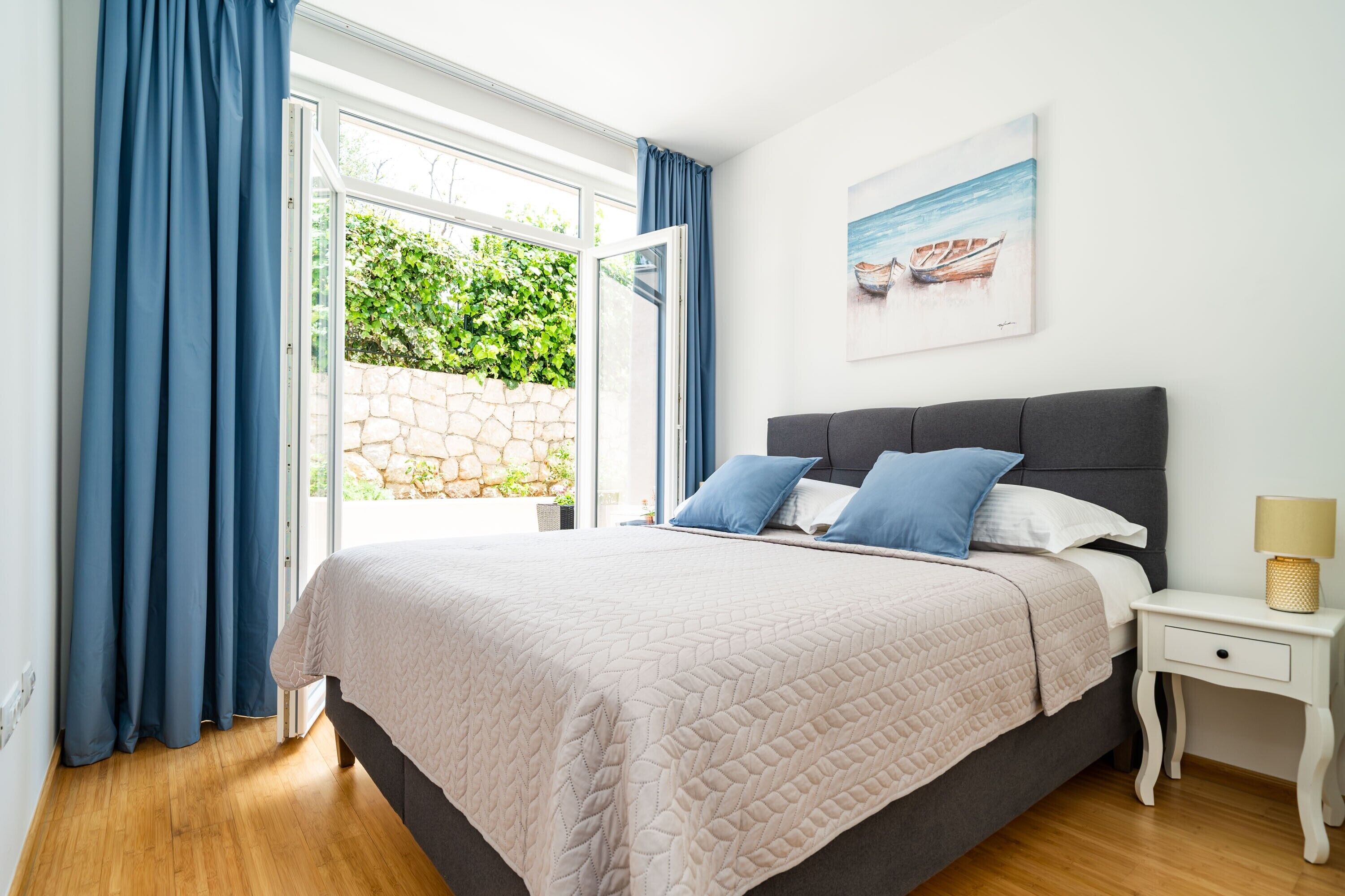 A bedroom with a bed, a nightstand, a lamp, a painting, and a view of greenery through a window.