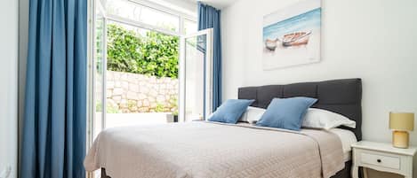 A bedroom with a bed, a nightstand, a lamp, a painting, and a view of greenery through a window.