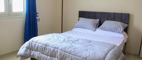 A neatly made bed with a white duvet and pillows, a dark headboard, and a blue curtain partially drawn to the side.