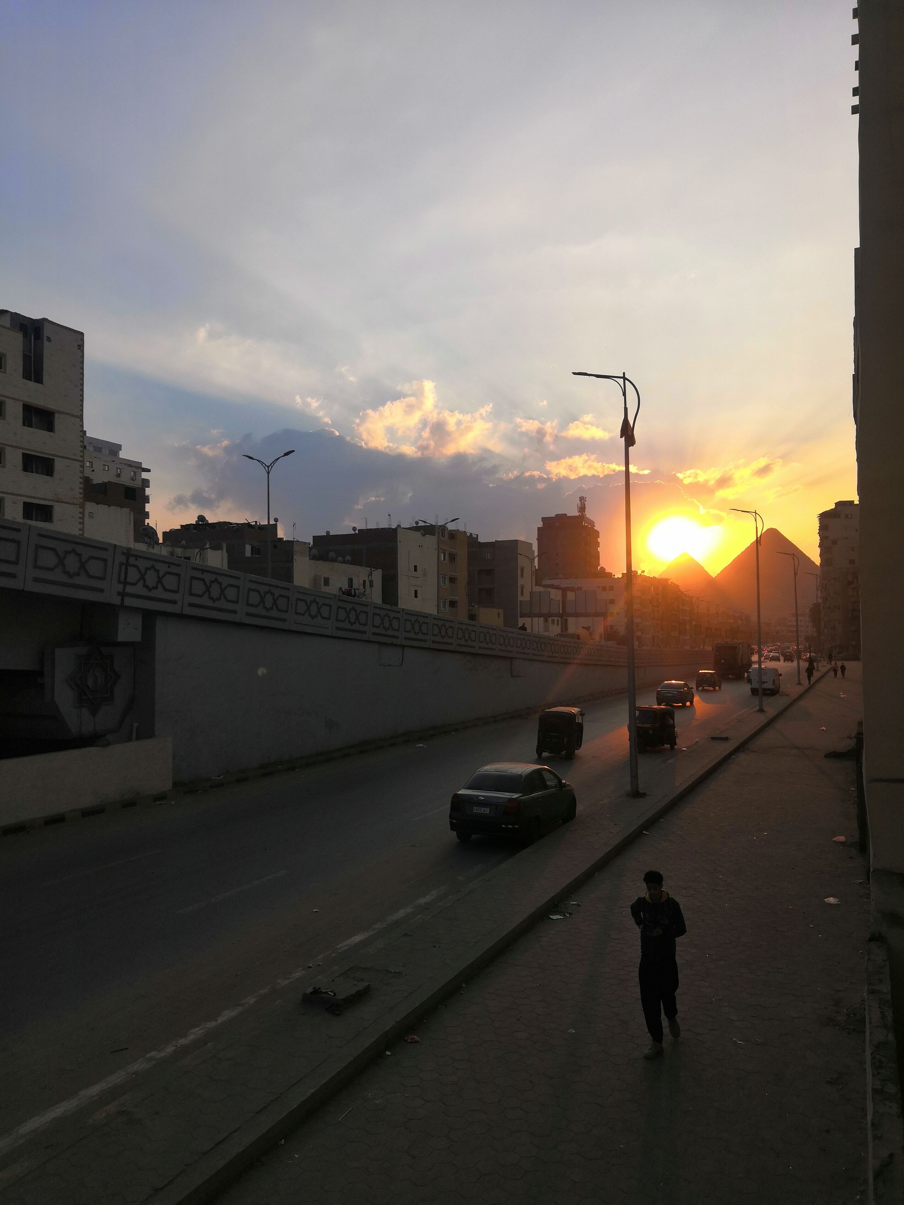 A city street at sunset with buildings, vehicles, and a pedestrian.