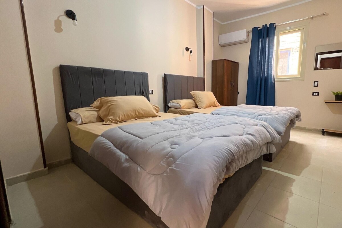 A bedroom with a bed, two bedside tables, a wardrobe, a window with curtains, an air conditioning unit, and a small plant on a shelf.