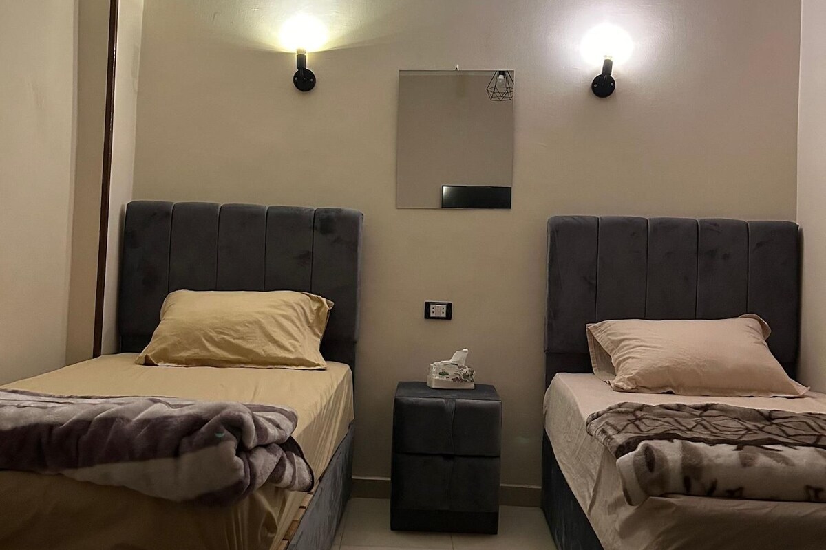 A bedroom with two beds, each with a headboard, a bedside table, and a wall-mounted light fixture.