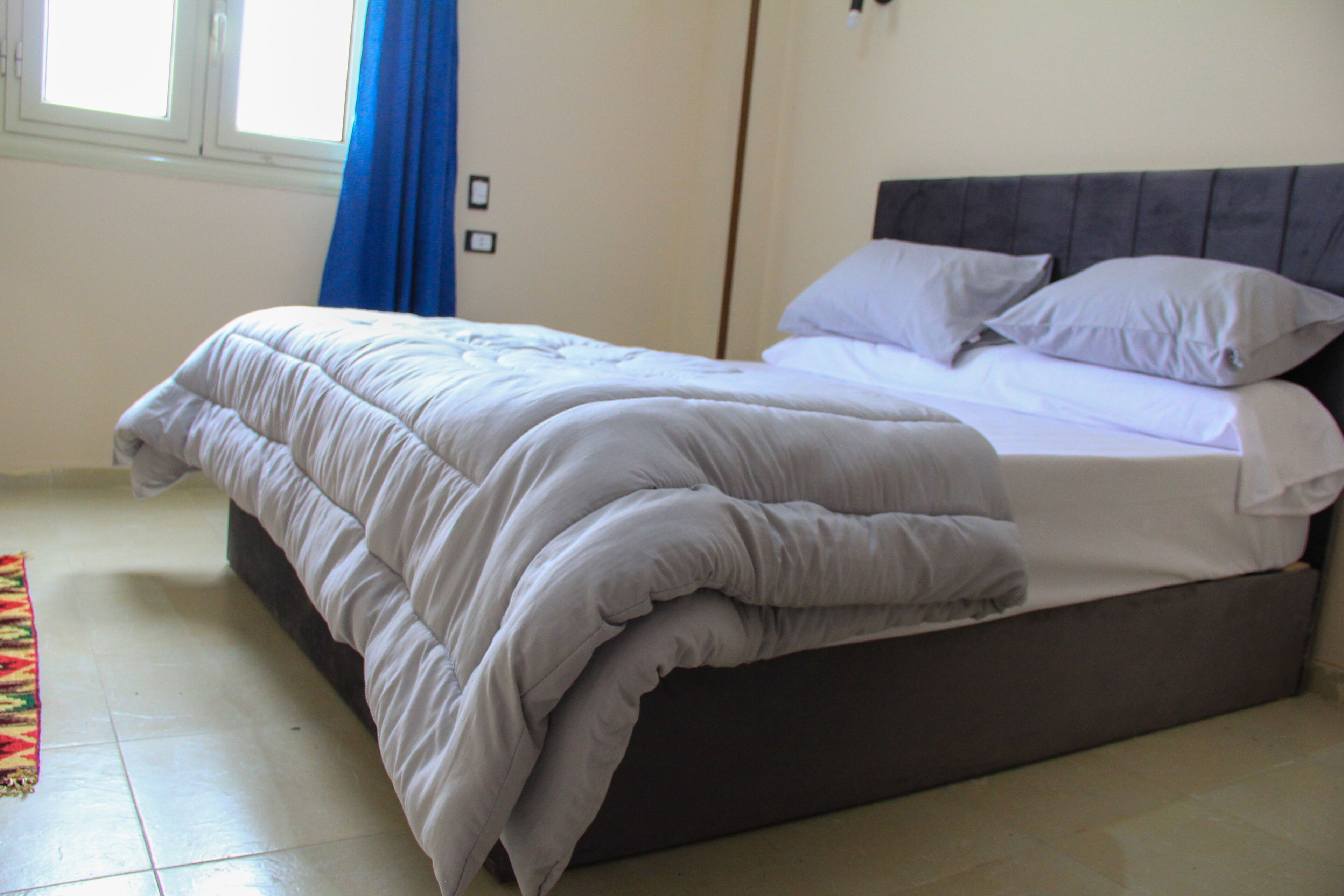 A neatly made bed with a grey headboard and white bedding in a room with a tiled floor and a window with blue curtains.