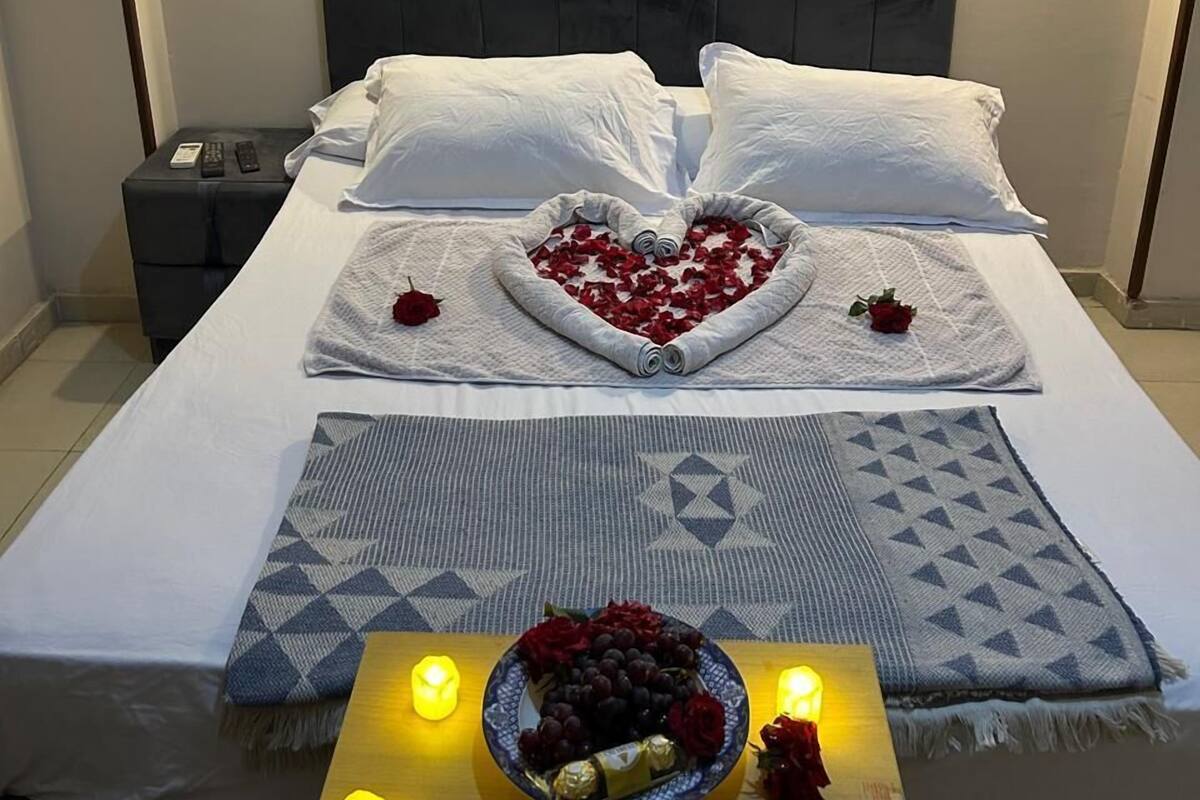A neatly made bed with a heart-shaped arrangement of rose petals and a small bouquet of roses on the bedside table.