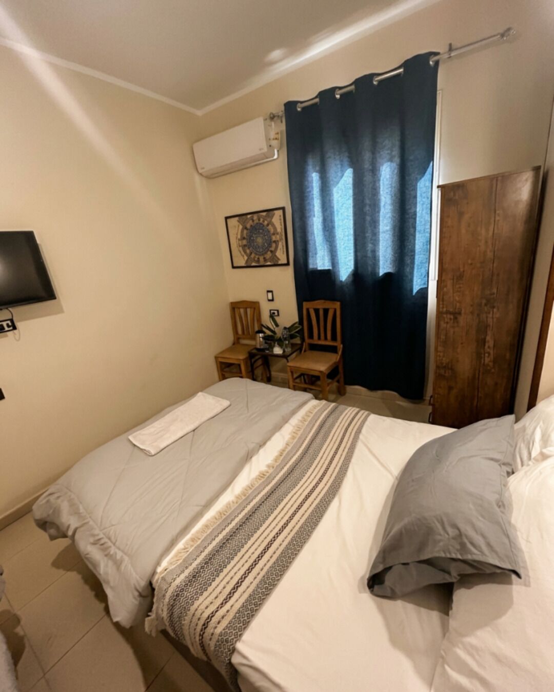 A bedroom with a bed, a TV, a desk with a chair, a wooden wardrobe, and a window with curtains.