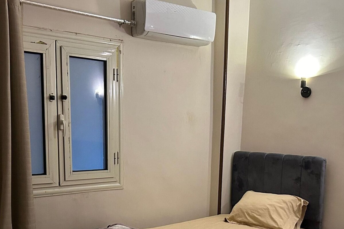 A bedroom with a bed, a bedside table, an air conditioner, a window with curtains, and a wall-mounted light.