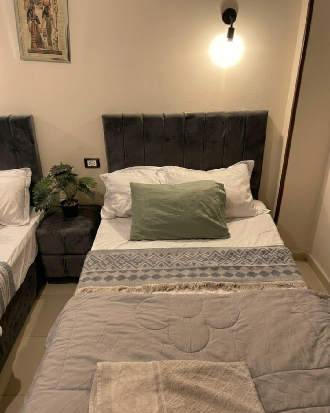 A bedroom with a bed, two nightstands, a potted plant, and a wall-mounted light fixture.