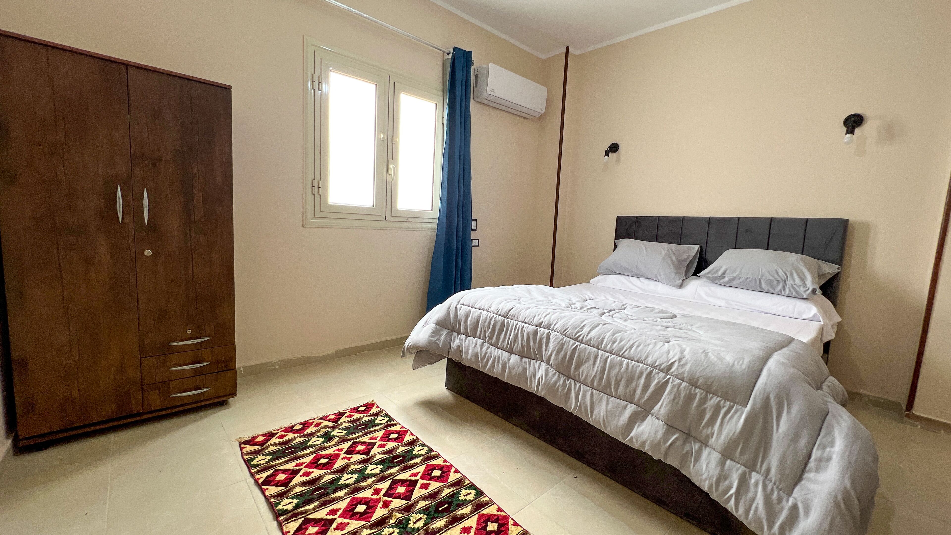 A bedroom with a bed, a wardrobe, a window with curtains, and an air conditioning unit.