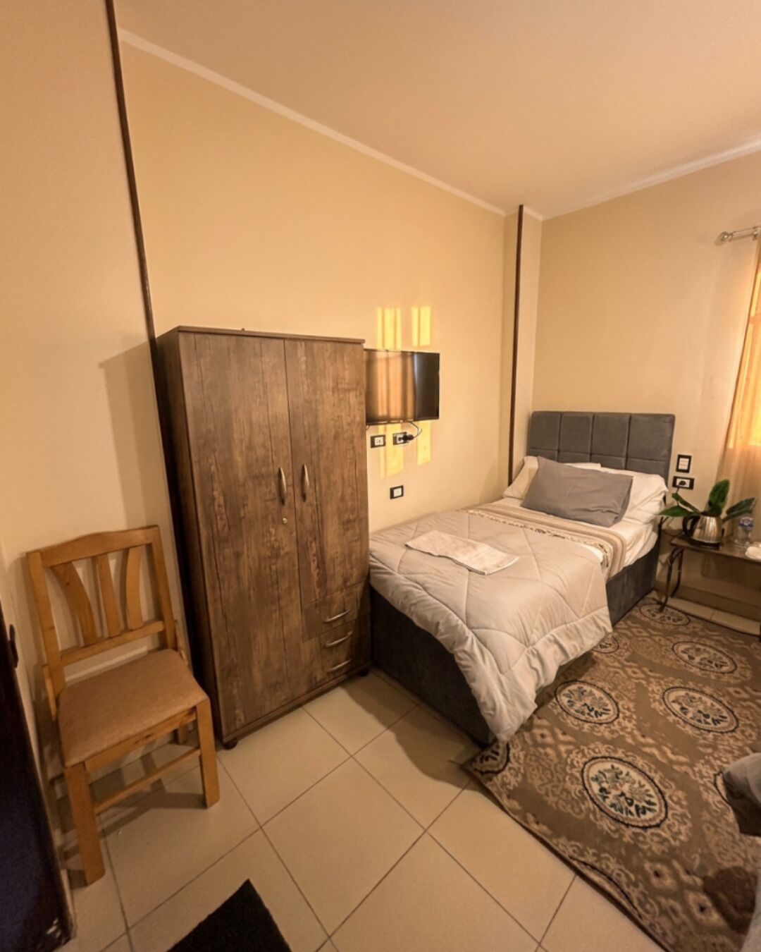 A bedroom with a bed, a wooden wardrobe, a chair, a bedside table with a lamp, and a patterned rug.