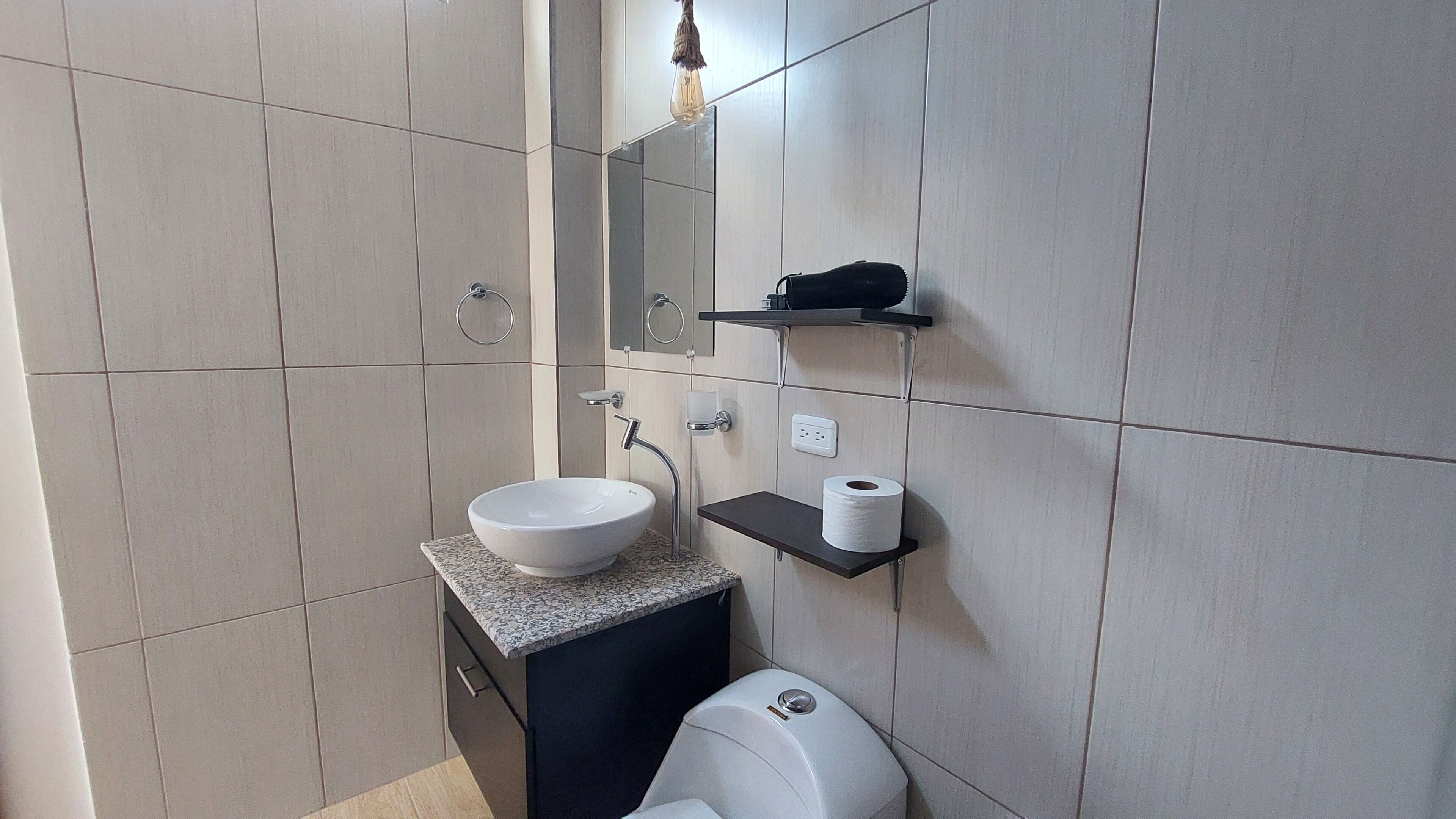Un baño moderno con un lavabo blanco, un inodoro, un sujetarropas, un perchero, un espejo y una repisa con un rollo de papel higiénico.