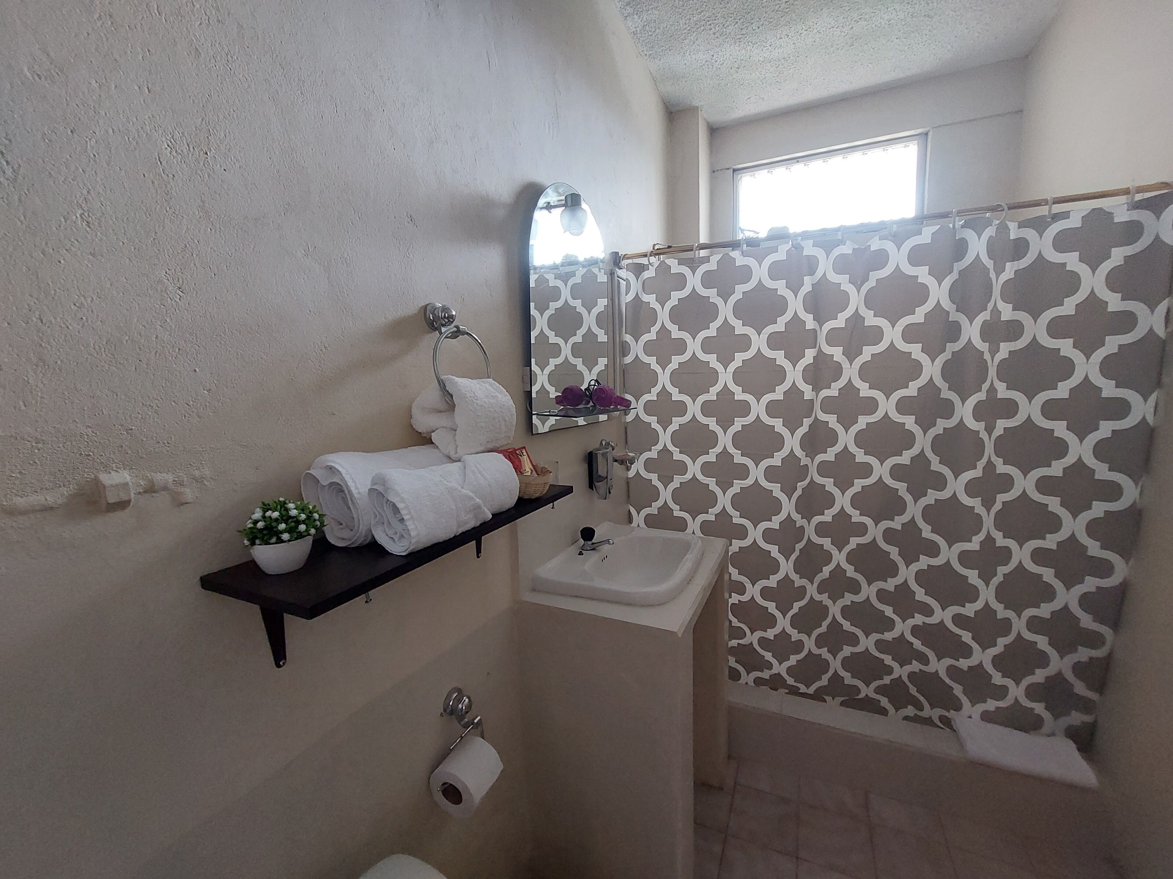 A bathroom with a patterned shower curtain, a sink, a toilet, and a shelf with towels and a potted plant.