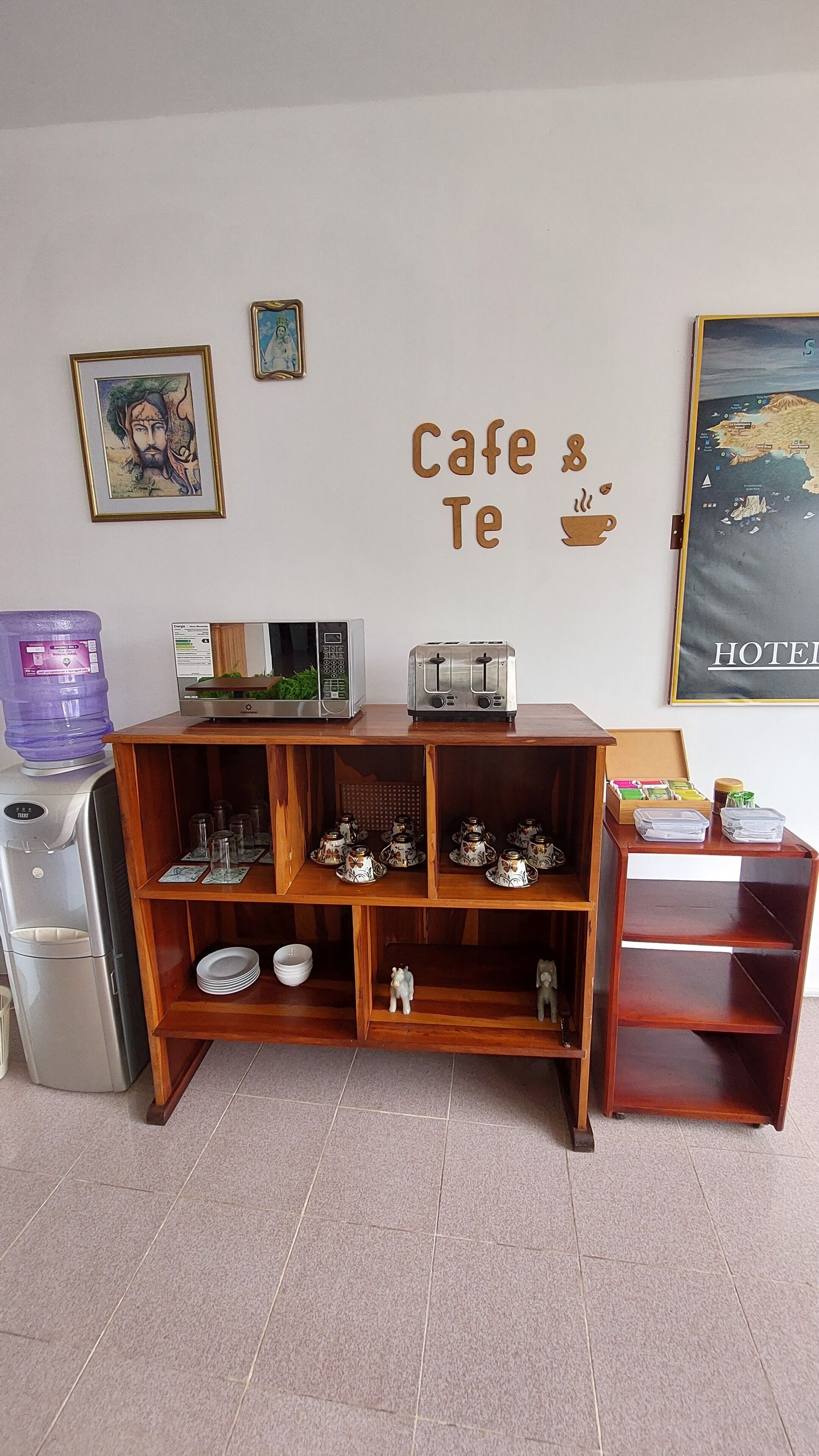 Una cuina americana amb nevera, microones i torradora. Hi ha prestatges amb tasses i plats, i un dispensador d'aigua. A la paret hi ha un cartell de 'Cafe & Te' i un mapa.