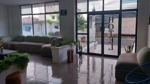 A hotel lobby with a sofa, chairs, a reception desk, and large windows with a view of the street and buildings. - Hotel Islas Galapagos (Puerto Baquerizo Moreno)