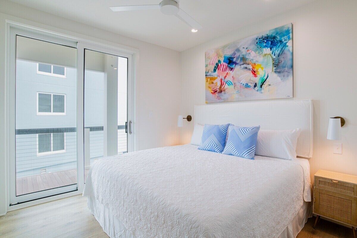 A bedroom with a large bed, a ceiling fan, a sliding glass door to a balcony, and a colorful abstract painting on the wall.
