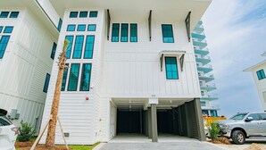 Un immeuble résidentiel moderne à plusieurs étages, doté d’une entrée de garage couverte, d’un palmier et d’un VUS garé.