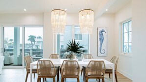 Un coin repas avec une table blanche et des chaises en osier, de grandes fenêtres et une œuvre d’art représentant un hippocampe accrochée au mur.
