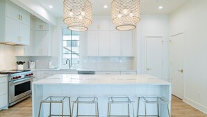Une cuisine moderne avec des meubles blancs, un grand îlot central doté de tabourets, des appareils en acier inoxydable et deux lustres en cristal suspendus.