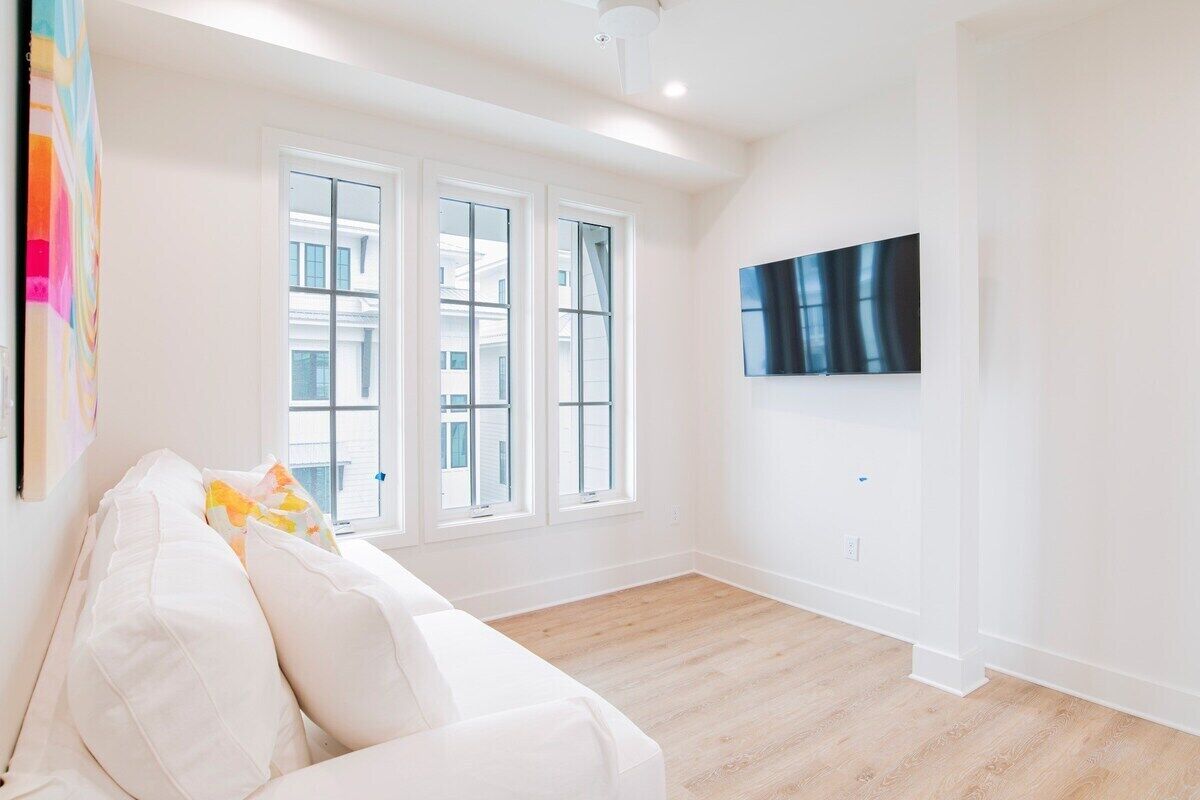 A spacious room with a white sofa, large windows, and a flat-screen TV mounted on the wall.