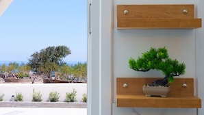 Quarto com prateleira de madeira exibindo uma árvore bonsai e um pequeno vaso, janela com vista para árvores e para o mar, e porta branca.