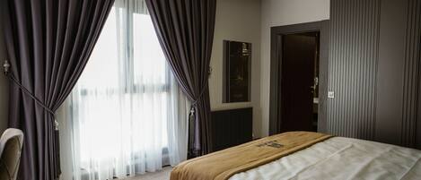Quarto de hotel com cama, uma cadeira, janela com cortinas, porta e radiador.