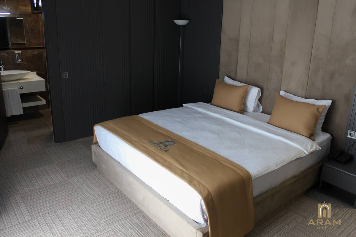 A hotel room with a large bed, a bedside table, a nightstand, a lamp, a bathroom with a sink and mirror, and a towel rack.
