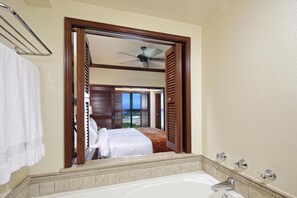 Um banheiro com banheira, chuveiro e um espelho refletindo um quarto com cama e janela.