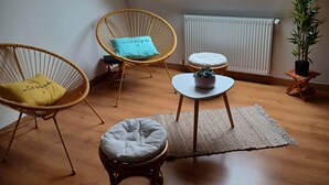 Ein Zimmer mit schrägen Wänden, einem Oberlicht, einem Heizkörper, einem kleinen runden Tisch, zwei Korbstühlen, einem runden Hocker und einer Topfpflanze.