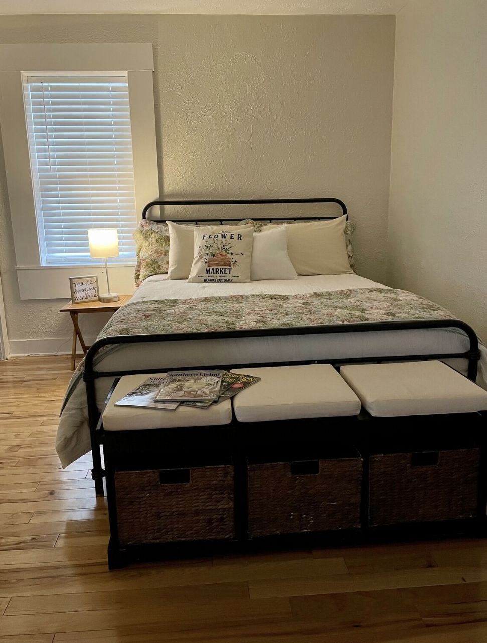 A bedroom with a metal bed frame, a bedside table, a lamp, and a window with blinds.