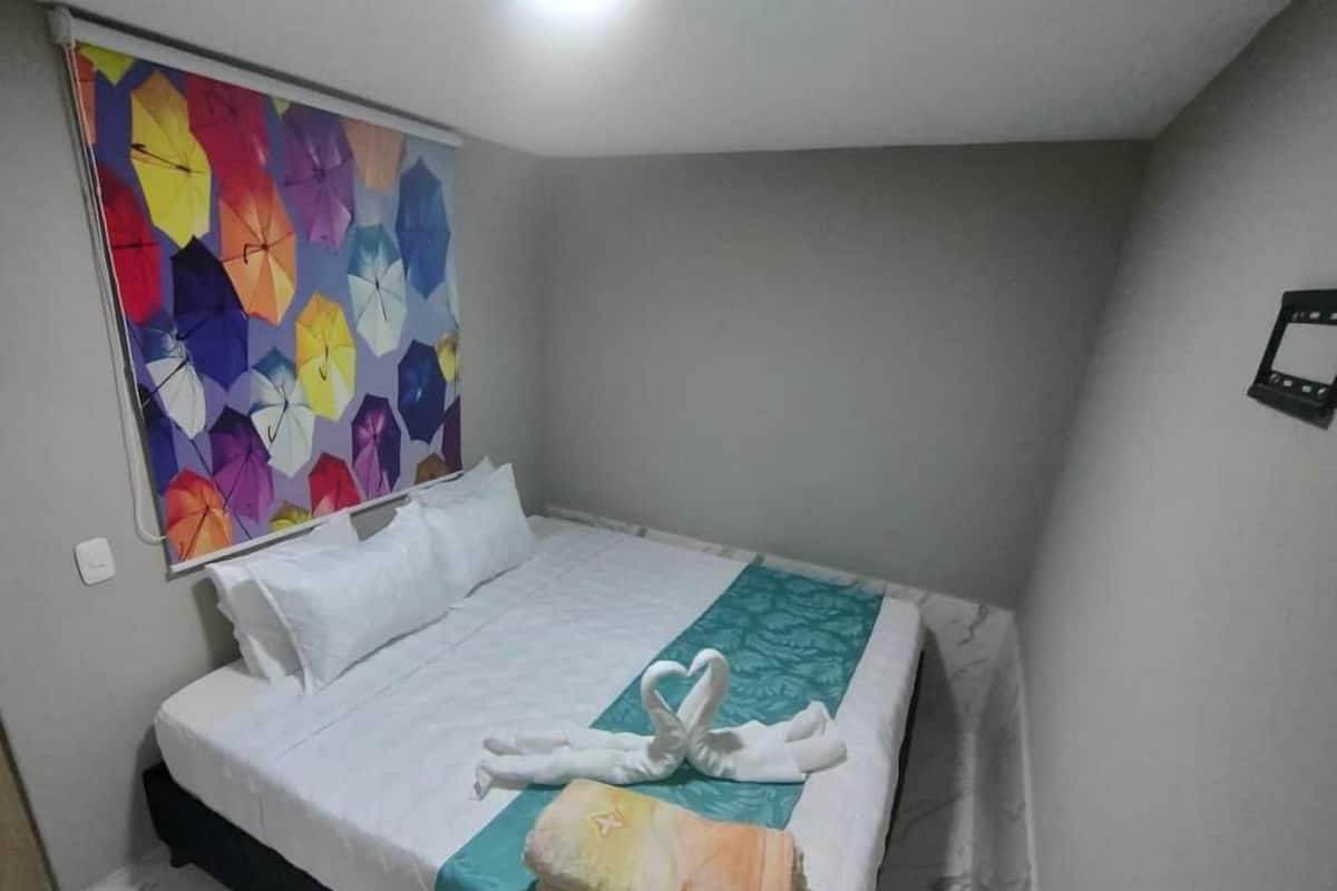 A hotel room with a bed, white linens, a teal bedspread, and a colorful umbrella artwork on the wall.