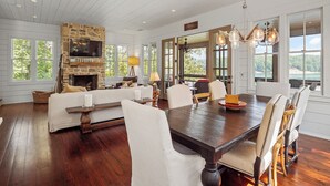 A spacious living room with a stone fireplace, large windows offering a view of the outdoors, and a dining area with a wooden table and chairs. - Central Perk - pet friendly, luxury cabin feel,  coffee! (Crane Hill)