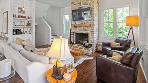 A spacious living room with a stone fireplace, a large TV mounted above it, and a cozy seating area with leather chairs and a sofa. - Central Perk - pet friendly, luxury cabin feel,  coffee! (Crane Hill)