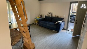 A loft-style room with a sloped ceiling, a ceiling fan, a large window, a blue sofa, a wooden ladder, and a dining area with a table and chairs. - Tiny House (not on water front) near lake martin, and boat ramp (Dadeville)