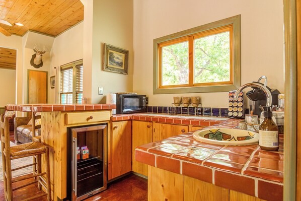 Una cocina con armarios de madera, microondas, refrigerador y ventana con vista a árboles.