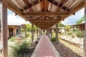 A covered walkway with wooden beams and a brick path, surrounded by various plants and small buildings. - Mucho Gusto in Bandera (Bandera)
