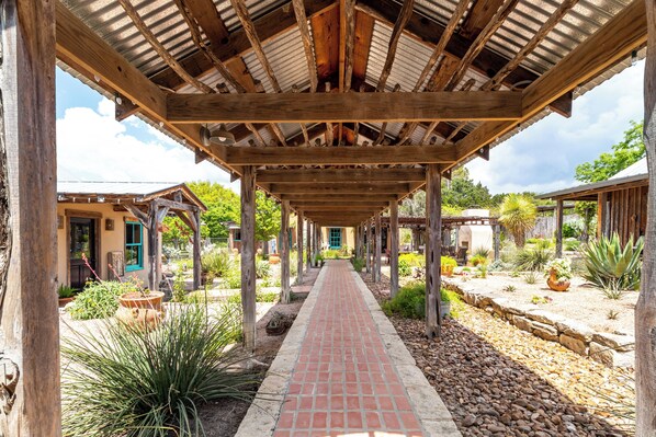 A covered walkway with wooden beams and a brick path, surrounded by various plants and small buildings. - Mucho Gusto in Bandera (Bandera)