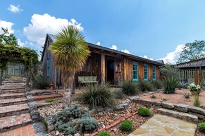 Un edificio rústico de madera con un porche, rodeado de un jardín ajardinado con diversas plantas y un sendero de piedra.