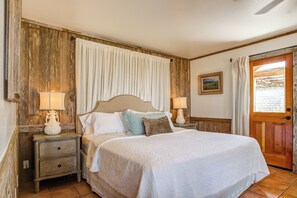 Un dormitorio con cabecera de madera, dos mesitas de noche con lámparas, una cama con colcha blanca y una puerta que ofrece vistas al exterior.