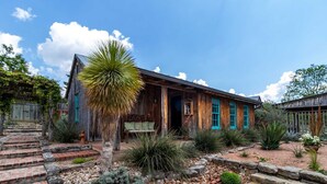 A rustic wooden building with a porch, surrounded by a garden with various plants and a stone pathway. - Mucho Gusto in Bandera (Bandera)