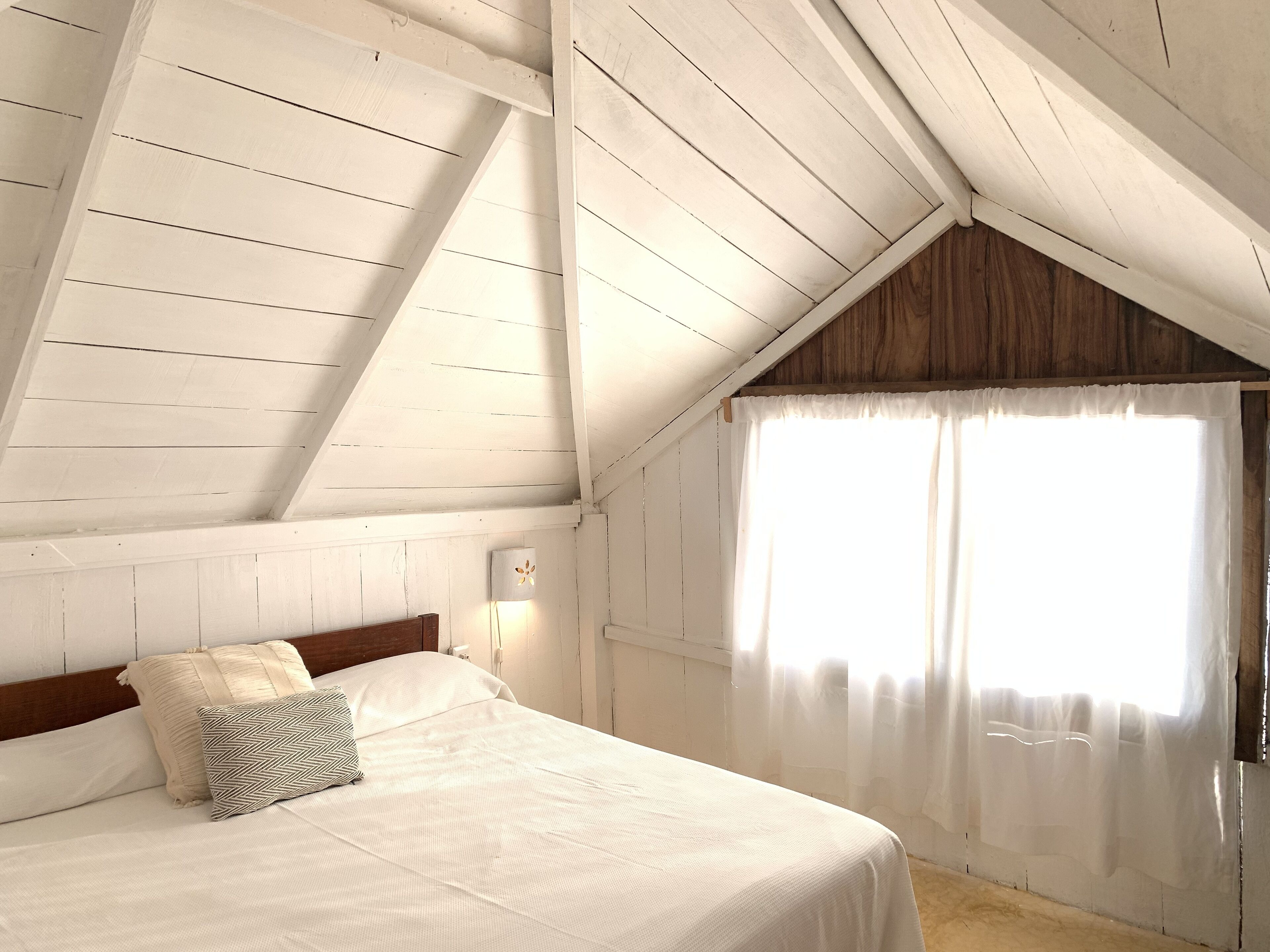 Une chambre avec un plafond en pente, un lit recouvert de draps blancs, deux oreillers, une lampe fixée au mur et une fenêtre ornée de rideaux légers.