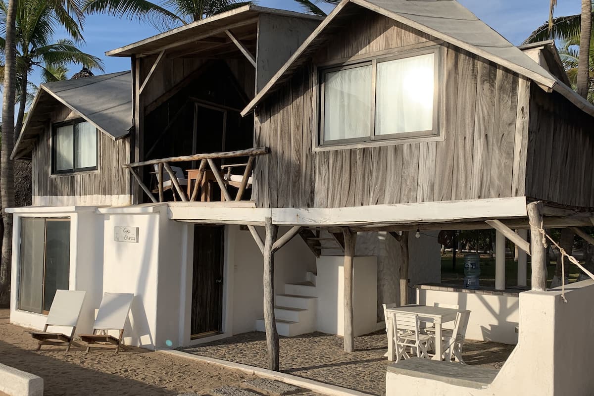 A wooden beach house with a balcony, a covered outdoor seating area, and a sandy ground.